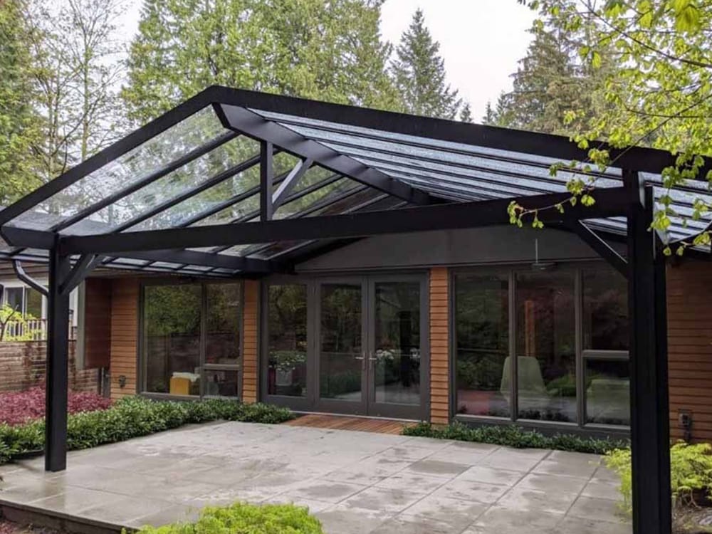 Sloped glass pergola in backyard Contemporary backyard pergola with sloped transparent roof panels and dark metal framing attached to a wooden exterior home.