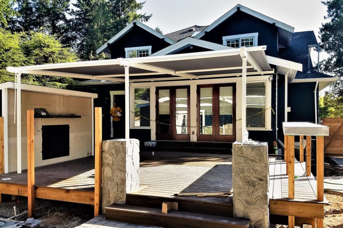 Backyard patio renovation featuring a wooden deck, stone pillars, retractable roof cover, and built-in outdoor fireplace in front of a modern black house.